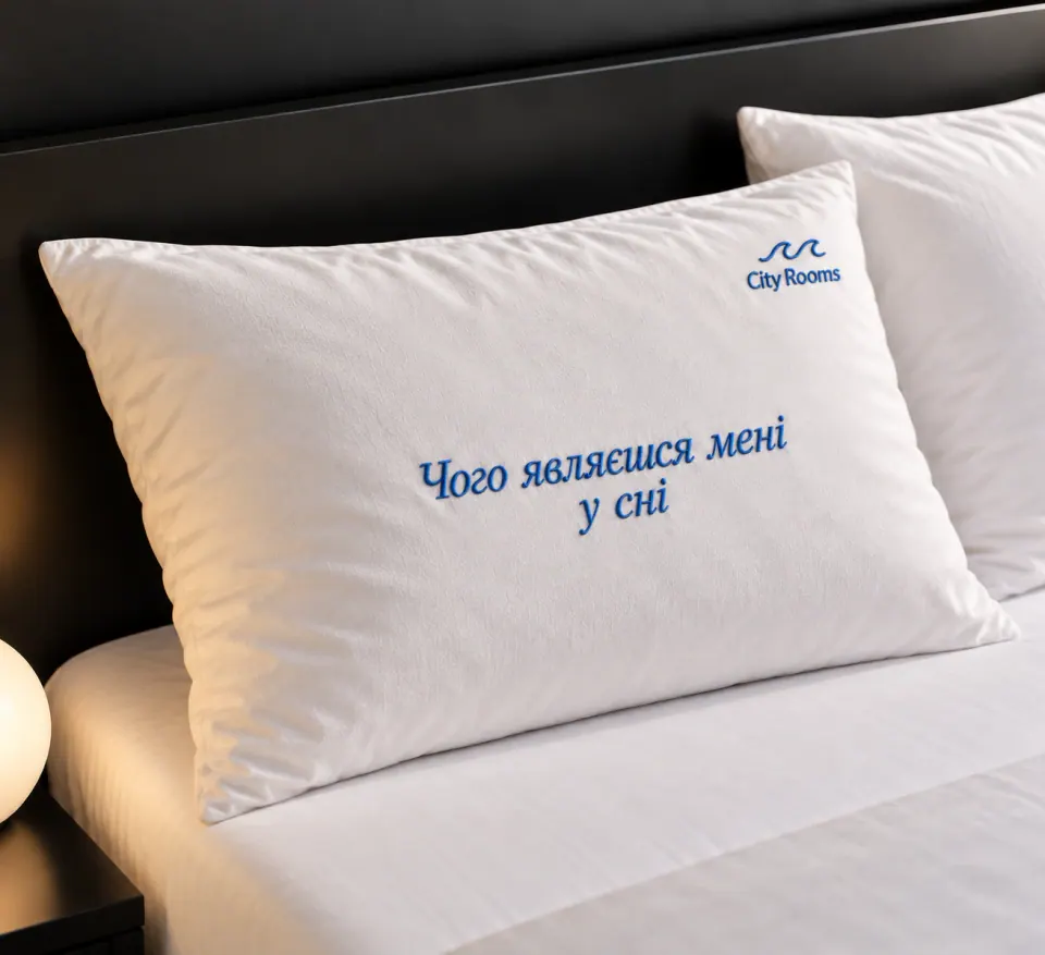 Синя вишивка фрази «Чого являєшся мені у сні» і логотипа City Rooms на білій готельній подушці